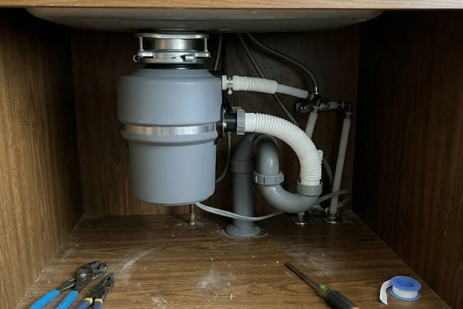 Inside a sink cabinet, a gray garbage disposal mounted under the sink with white flexible drain hoses and a gray P-trap, with pliers, a screwdriver and thread tape on the cabinet floor