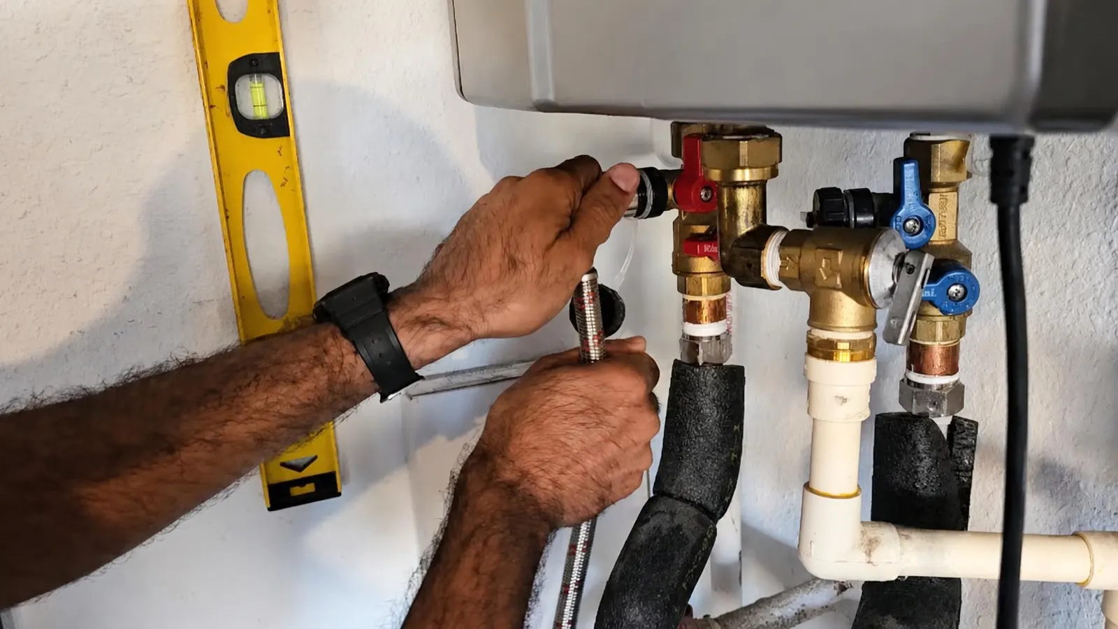 Hands connecting a flexible hose to brass shutoff valves and a relief valve beneath a wall-mounted tankless water heater, with a yellow level on the wall